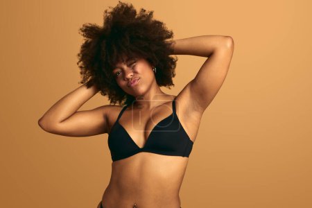 Self assured young plus size African American female model in black lingerie winking and looking at camera, while touching curly dark hair against beige background