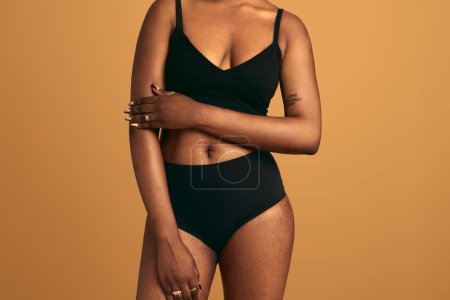 Photo for Anonymous African American plump female model in black underwear keeping hand on arm while standing against beige background in studio - Royalty Free Image