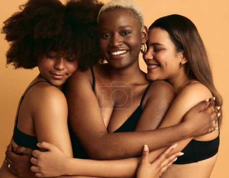 Téléchargez les photos : Copines multiraciales souriantes en lingerie noire s'embrassant tout en se tenant debout sur fond orange concept de convivialité et de soutien - en image libre de droit