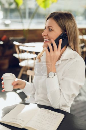 Photo for Positive adult female copywriter sitting at round table near notebook with notes and drinking coffee from paper cup, while talking on phone and looking away - Royalty Free Image
