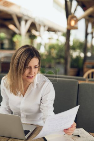 Photo for Concentrated woman in white shirt reading text on paper while working remotely on laptop in outdoor cafe - Royalty Free Image