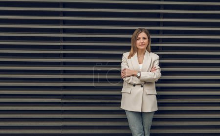 Téléchargez les photos : Femme d'affaires respectable en vêtements décontractés intelligents debout avec les mains croisées contre le mur rayé et regardant la caméra - en image libre de droit