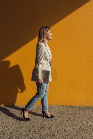 Foto de Cuerpo completo de mujer de negocios en chaqueta de moda y jeans que sostienen el ordenador portátil mientras pasa por la pared amarilla en la calle de la ciudad bajo el sol - Imagen libre de derechos
