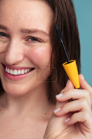 Photo for Portrait of crop cheerful female model with mascara applicator looking at camera and smiling while doing eye makeup against blue background - Royalty Free Image