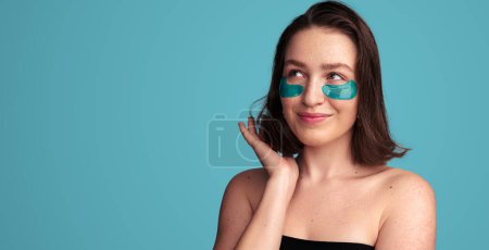 Téléchargez les photos : Contenu jeune modèle féminin avec bandeaux oculaires et épaules nues souriant et regardant loin tout en touchant les cheveux foncés sur fond bleu - en image libre de droit