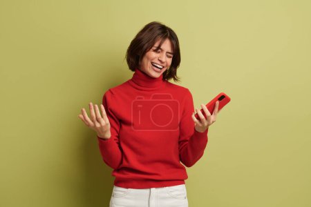 Photo for Delighted young female millennial with dark hair in red turtleneck laughing with closed eyes while messaging on mobile phone against green background - Royalty Free Image