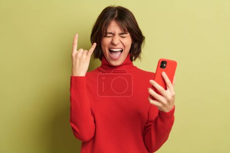 Foto de Alegre joven hembra en suéter rojo sonriendo y mostrando dos dedos gesto mientras sostiene un teléfono inteligente sobre fondo verde - Imagen libre de derechos