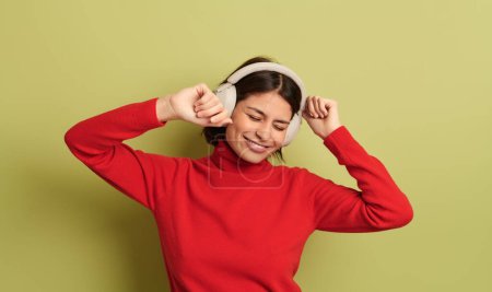 Foto de Mujer hispana joven excitada con el pelo oscuro en cuello alto rojo sonriendo con los ojos cerrados, mientras escucha música en auriculares inalámbricos y bailando sobre fondo rojo - Imagen libre de derechos