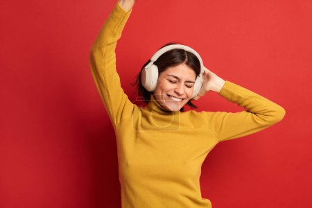 Photo for Delighted young female millennial with dark hair in yellow turtleneck smiling happily with closed eyes, while listening to favorite song in wireless headphones and dancing against red background - Royalty Free Image