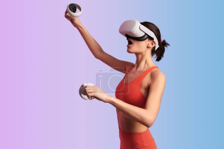 Photo for Side view of young female in VR goggles and with controllers playing video game against gradient background - Royalty Free Image