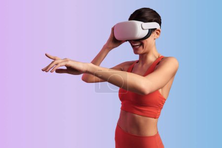 Young cheerful female touching VR goggles while exploring cyberspace laughing and pointing away watching funny video against gradient background