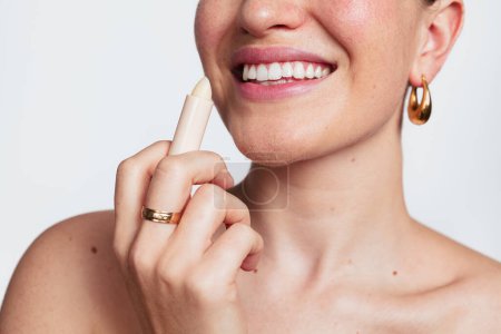 Foto de Feliz señora anónima con hombros desnudos con pendientes y anillo mientras se aplica lápiz labial higiénico sobre fondo blanco - Imagen libre de derechos