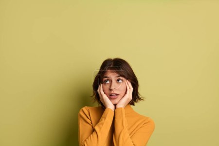 Photo for Thoughtful young Hispanic female model with dark hair in bright yellow sweater touching cheeks and looking up dreamily against green background - Royalty Free Image