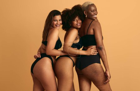 Téléchargez les photos : Jeunes copines multiraciales souriantes portant des sous-vêtements noirs debout près de l'autre taille touchante sur fond orange concept de fraternité - en image libre de droit