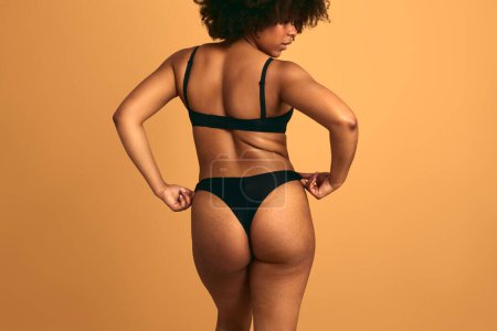 Foto de Vista posterior del modelo femenino africano más tamaño tocando las bragas mientras está de pie sobre fondo naranja en el estudio - Imagen libre de derechos