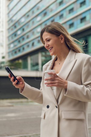Téléchargez les photos : Positif jeune femme dans blazer élégant navigation téléphone mobile tout en marchant sur la rue de la ville - en image libre de droit