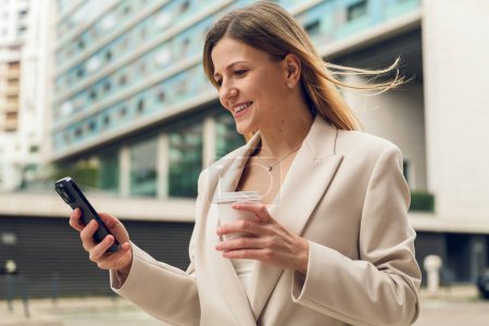 Photo for Self assured adult happy female entrepreneur with long blond hair in stylish suit, reading message on smartphone and smiling while walking on street with cup of takeaway coffee during break - Royalty Free Image