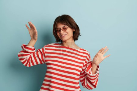 Foto de Mujer hispana joven y segura de sí misma con cabello castaño en suéter rojo y blanco rayado y gafas de sol de moda, sonriendo con los ojos cerrados mientras baila sobre fondo azul - Imagen libre de derechos