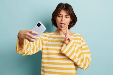 Téléchargez les photos : Jeune femme positive en pull jaune prenant autoportrait sur smartphone et montrant la langue avec signe de paix sur fond bleu - en image libre de droit
