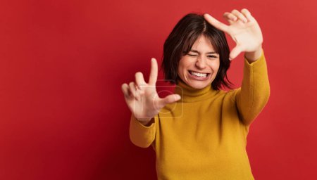 Téléchargez les photos : Joyeux jeune femme au col roulé jaune pull souriant tout en montrant geste photo avec les yeux fermés sur fond rouge - en image libre de droit