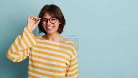 Téléchargez les photos : Jeune femme positive en chemise rayée et lunettes souriantes et regardant la caméra tout en se tenant debout sur fond bleu avec espace de copie - en image libre de droit