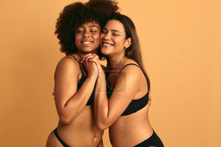 Photo for Cheerful young multiracial female models in black underwear embracing each other and smiling with closed eyes, while standing close and holding hands against beige background together in studio - Royalty Free Image