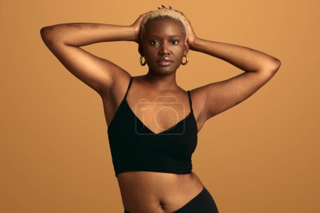 Foto de Joven modelo femenino afroamericano con pelo rubio corto con ropa interior negra tocando la cabeza mientras mira la cámara contra el fondo naranja - Imagen libre de derechos
