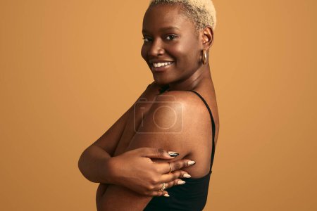 Téléchargez les photos : Vue latérale du modèle féminin afro-américain joyeux jeune en soutien-gorge noir regardant la caméra et souriant sur fond beige avec les mains sur la poitrine - en image libre de droit