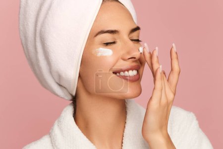 Positive young female model with perfect skin and towel on head smiling with closed eyes, while applying moisturizing cream on cheeks and nose after bath against pink background