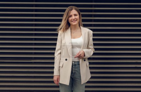 Téléchargez les photos : Femme d'affaires réussie positive en veste beige et jeans debout contre porte de garage rayé regardant la caméra - en image libre de droit