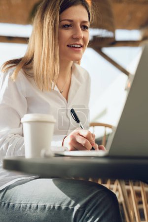 Téléchargez les photos : Femme adulte souriante en chemise blanche et jeans assis à table avec café dans une tasse en papier et des notes d'écriture, tout en travaillant à distance sur un ordinateur portable à partir d'un café - en image libre de droit