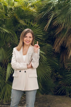 Téléchargez les photos : Jeune femme d'affaires positive en tenue décontractée intelligente debout contre les plantes vertes exotiques et pointant du doigt vers le haut en regardant la caméra - en image libre de droit