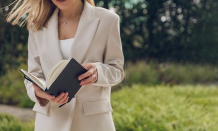 Photo for Crop unrecognizable female in white blazer standing on grassy field near trees and reading interesting book - Royalty Free Image