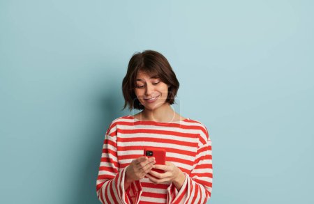 Foto de Joven mujer hispana positiva millennial con cabello oscuro en ropa casual sonriendo felizmente mientras mensajea en el teléfono móvil contra fondo azul - Imagen libre de derechos