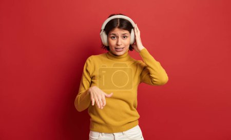 Foto de Confiada joven étnica milenaria con cabello oscuro en cuello alto amarillo escuchando música en auriculares inalámbricos y mirando a la cámara, mientras finge ser DJ contra el fondo rojo - Imagen libre de derechos