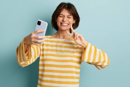 Foto de Joven mujer alegre étnica en camisa a rayas sonriendo mientras toma autorretrato en el teléfono inteligente, sobre fondo azul - Imagen libre de derechos
