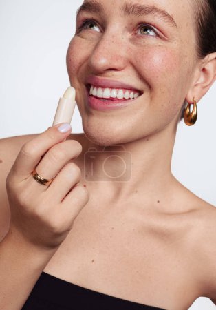 Téléchargez les photos : Crop modèle féminin confiant positif avec taches de rousseur et les épaules nues souriant et détournant les yeux tout en appliquant le baume à lèvres sur fond blanc - en image libre de droit