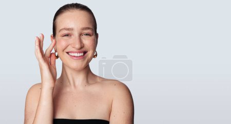 Téléchargez les photos : Jeune femme féminine aux cheveux foncés et aux épaules nues touchant le visage et souriant à la caméra sur fond gris - en image libre de droit