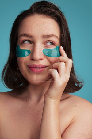 Photo for Young attractive female model touching eye patch while enjoying skincare treatment against turquoise background looking up - Royalty Free Image