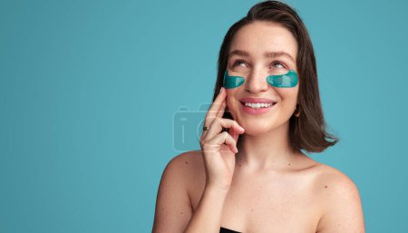 Téléchargez les photos : Joyeux jeune femme appliquant des patchs hydrogel bleu sur le visage tout en se tenant debout sur fond bleu et en levant les yeux - en image libre de droit