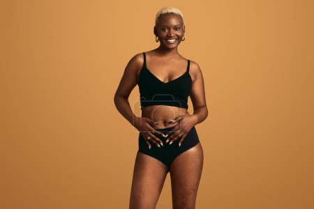 Foto de Joven modelo femenina afroamericana alegre en lencería negra con pelo corto mirando a la cámara y sonriendo, sobre fondo beige mientras toca el vientre en el estudio - Imagen libre de derechos