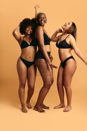 Photo for Full body of happy young multiethnic plus size girlfriends wearing black underwear, dancing in studio against orange background with raised arms while having fun together - Royalty Free Image