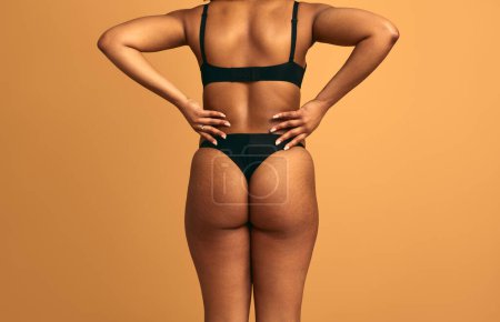 Photo for Back view of crop unrecognizable African American plus size female model in black underwear standing against beige background background with hands on waist - Royalty Free Image
