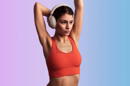 Photo for Fit young female athlete in red sports top and headphones listening to music while stretching arms during workout against gradient background - Royalty Free Image