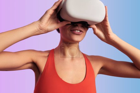 Téléchargez les photos : Jeune femme en haut rouge touchant lunettes VR tout en explorant le monde virtuel jouer jeu vidéo sur fond de dégradé - en image libre de droit