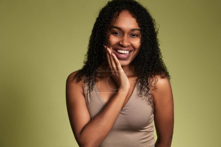 Photo for Young smiling African American woman with curly hair wearing top and caressing gentle skin of face looking at camera on green background - Royalty Free Image
