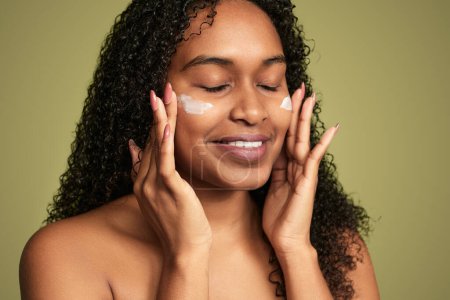 Cheerful African American female with curly hair applying moisturizing cream on face with closed eyes on green background
