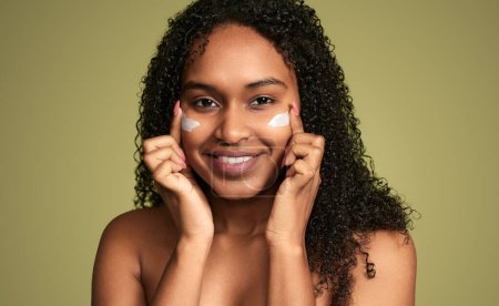 Foto de Contenido joven mujer afroamericana con la piel de la cara impecable untando crema bajo los ojos mientras mira la cámara en el fondo verde - Imagen libre de derechos