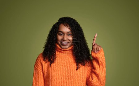 Téléchargez les photos : Femme afro-américaine joyeuse aux cheveux bouclés en pull orange souriant et regardant la caméra sur fond vert tout en pointant vers le haut - en image libre de droit