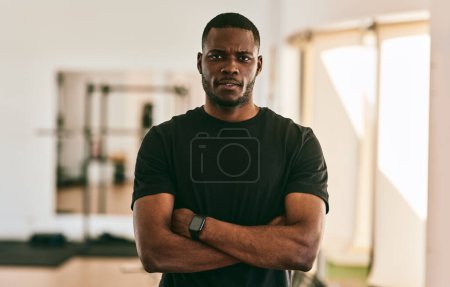 Photo for Self assured adult black male coach in t shirt with smart watch crossing hands while standing in gym against blurred background - Royalty Free Image